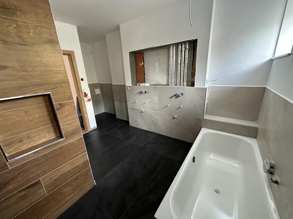 Modernes Badezimmer im Umbau mit weißer Badewanne, dunklen Bodenfliesen und Wandfliesen in Holzoptik.